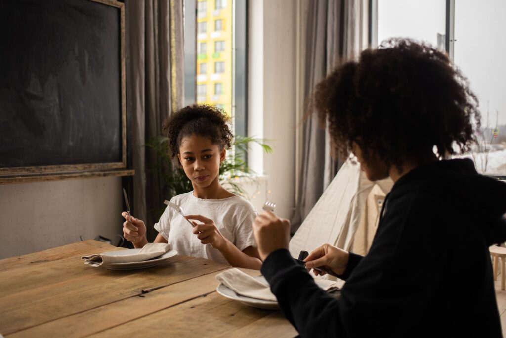 骰子吹牛玩法:你該注意的小技巧 3 Pensive African American girl sitting at wooden table with plate and cutlery in hands while learning dining etiquette with anonymous mother